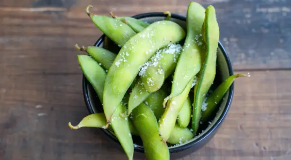 Grüne Edamame-Schoten, leicht gesalzen, in einer Schüssel auf Holzhintergrund. Was sind Edamame? Ein gesunder Snack!