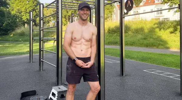Ein trainierter Mann steht lächelnd vor einer Calisthenics-Anlage im Freien.