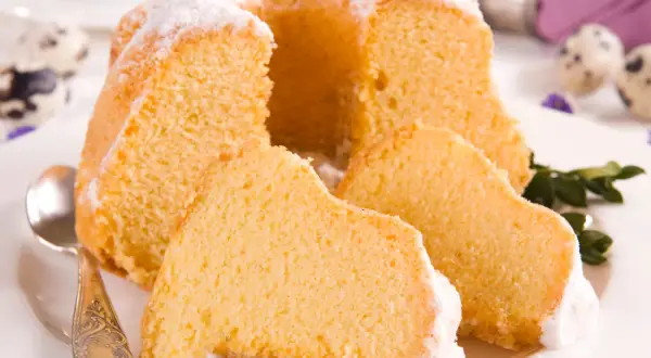 Puszysta babka z mascarpone, oprószona cukrem pudrem, z kilkoma kawałkami na talerzu. Obok leży łyżeczka.