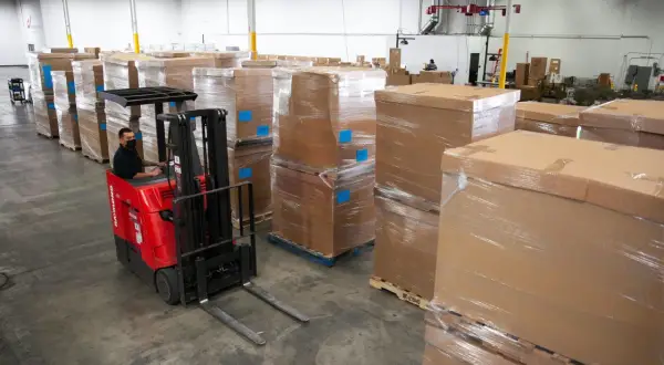 Magazyn z paletami towarów, pracownik w masce obsługuje wózek widłowy. Efektywne **cross docking** w logistyce.