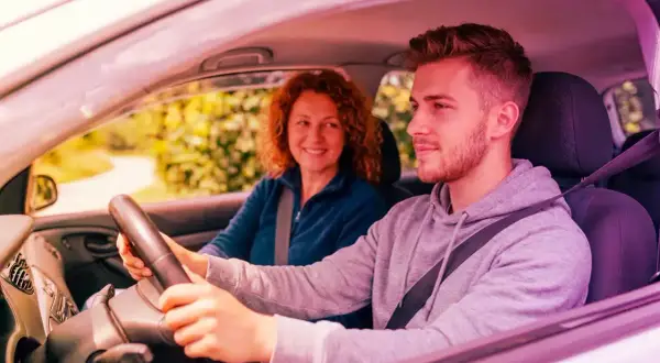 Giovane uomo guida, madre sorride. Ricorda che l'assicurazione auto deve essere intestata al proprietario per la massima tranquillità.