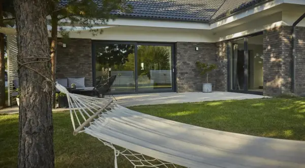 Hamak w ogrodzie nowoczesnego bungalow z ceglaną elewacją i dużymi przeszkleniami.