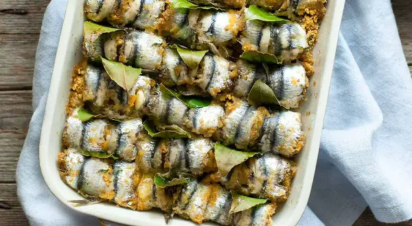Sarde a beccafico pronte per essere infornate, disposte in una teglia con foglie di alloro e panatura dorata.