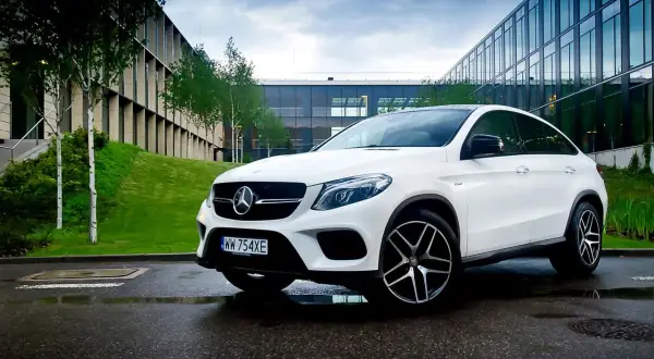 Biały Mercedes GLE 450 zaparkowany na mokrym asfalcie przed nowoczesnym budynkiem.