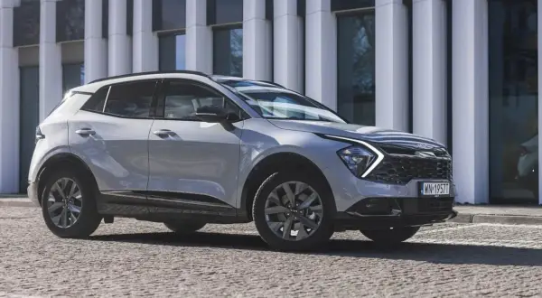 Nowy Kia Sportage Anniversary w jasnym kolorze, zaparkowany przed nowoczesnym budynkiem. Stylowy SUV na kamienistej nawierzchni.