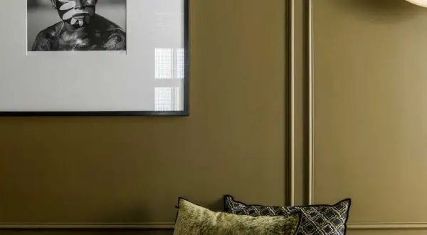 Un pan de mur vert olive, orné d'un portrait encadré d'un jeune homme au visage peint, et de coussins décoratifs.