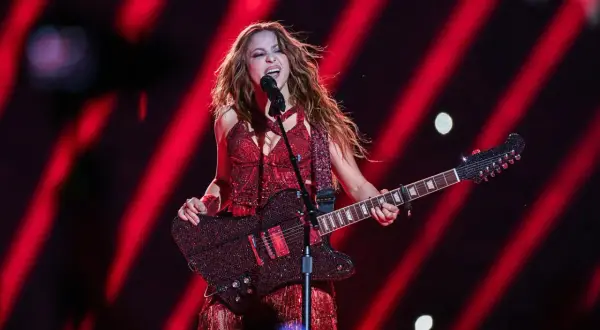 Shakira w czerwonym stroju śpiewa i gra na gitarze na tle czerwonych świateł.