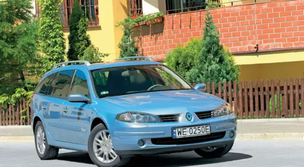 Błękitne kombi Renault Laguna 2 z relingami dachowymi stoi przed domem z żółtą elewacją i drewnianym płotem.
