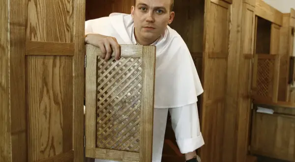 Ksiądz w białej albie otwiera kratkę konfesjonału, czekając, kiedy jest spowiedź w kościele.