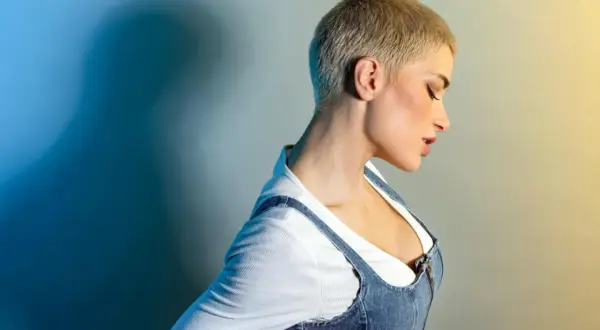 Kobieta z **buzz cut blond** w dżinsowej sukience na szelkach i białej bluzce. Ciemny cień na niebieskim tle.