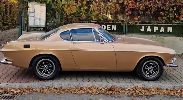 Klasyczny Volvo P1800 w kolorze złotym, zaparkowany na chodniku, z jesiennymi liśćmi wokół.