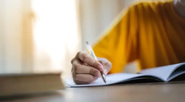 Main d'une personne écrivant dans un cahier, peut-être pour l'archive des noms de famille algériens.