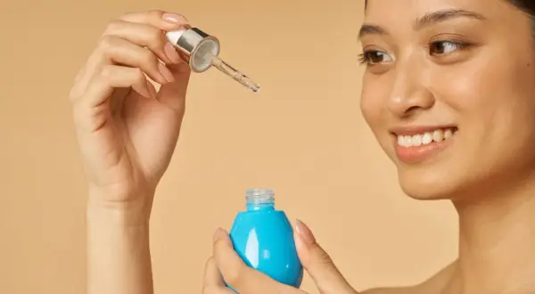 Uśmiechnięta kobieta trzymająca pipetkę z serum i niebieską buteleczkę. Poznaj the ordinary jak stosować, by uzyskać promienny blask.