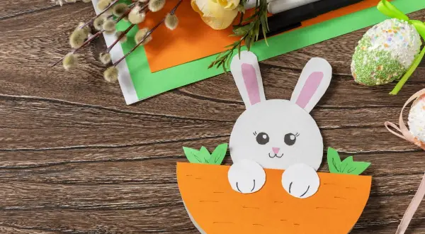 Uroczy króliczek z papieru kolorowego wychyla się z marchewki. Obok pisanki z papieru kolorowego, bazie i flamastry.