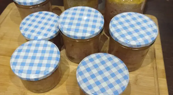 Mehrere Gläser mit selbstgemachter Leberwurst, verschlossen mit blau-weißen Karodeckeln, stehen auf einem Holzbrett.