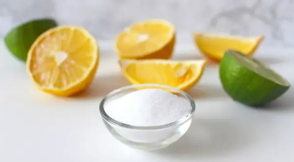 Una ciotola di cristalli bianchi, che ricordano la struttura dell'acido citrico, circondata da spicchi di limone e lime freschi.