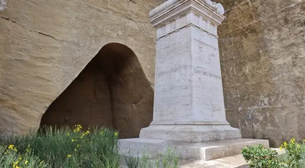 Monumento a Giacomo Leopardi nel Parco Virgiliano a Piedigrotta, con un'antica grotta sullo sfondo e fiori gialli.