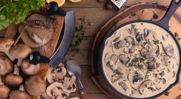 Cremige Rahmchampignons in einer Pfanne, daneben frische Champignons, Kräuter, Zitrone und Gewürze. Ein leckeres Rahmchampignons Rezept zum Nachkochen.