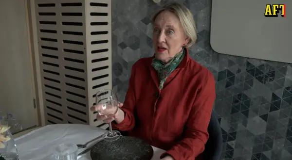 Äldre kvinna med röd skjorta och scarf håller ett glas vin. Hon sitter vid ett dukat bord, redo för en vett och etikett middag.