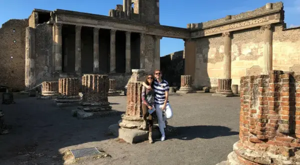 Pareja posa frente a ruinas antiguas, preguntándose donde está Pompeya. El sol ilumina columnas rotas y edificios históricos.