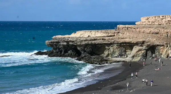 Fuerteventura Urlaub Tipps: Schwarzer Strand mit Felsen und türkisblauem Meer. Menschen spazieren und entspannen.