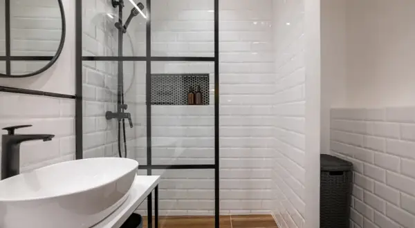Moderno murete baño con ducha de cristal, lavabo blanco y grifería negra. Azulejos blancos y suelo de madera.