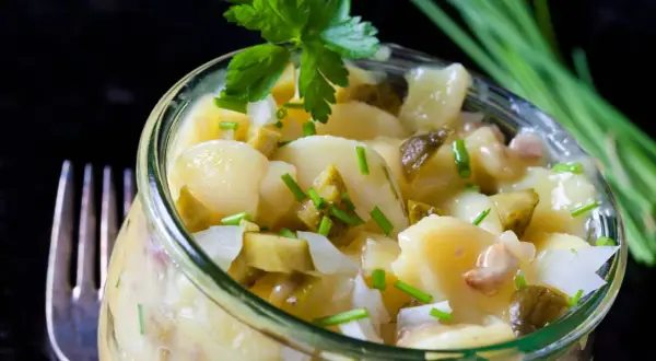 Ein köstlicher Kartoffelsalat ohne Mayo, mit Gurkenstücken und frischem Schnittlauch garniert, serviert in einem Glas.