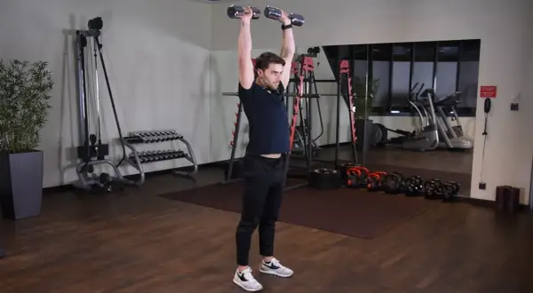 Wyciskanie hantli nad głowę angażuje mięśnie naramienne i triceps. Mężczyzna wykonuje ćwiczenie w siłowni.
