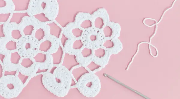 White Irish crochet lace with flowers and hearts, a crochet hook, and white yarn on a pink background.