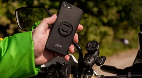 Dłoń trzyma czarne etui na telefon z mocowaniem SP Connect, obok uchwytu na kierownicy motocykla.