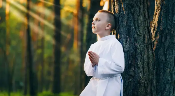 Chłopiec w albie modli się w lesie, promienie słońca przebijają się przez drzewa. Piękne sesje komunijne w plenerze.