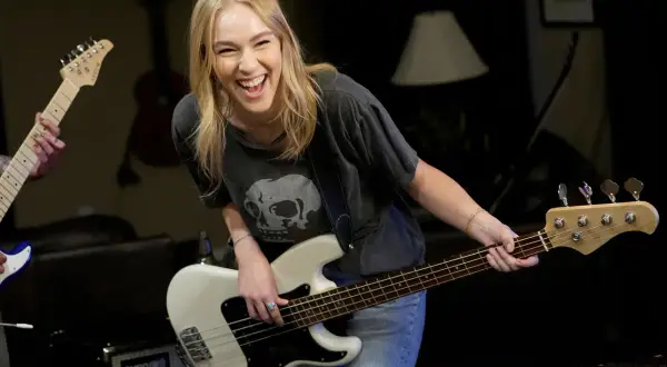 Uśmiechnięta blondynka gra na białym bas instrumencie, w tle inny gitarzysta.
