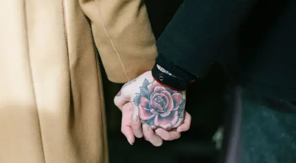 Manos entrelazadas, una con un tatuaje de rosa en fase lechosa, la otra envuelta en un abrigo camel.