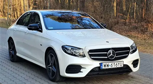 Biały Mercedes E300e z czarnym dachem i lusterkami, zaparkowany na brukowanej ścieżce w lesie.