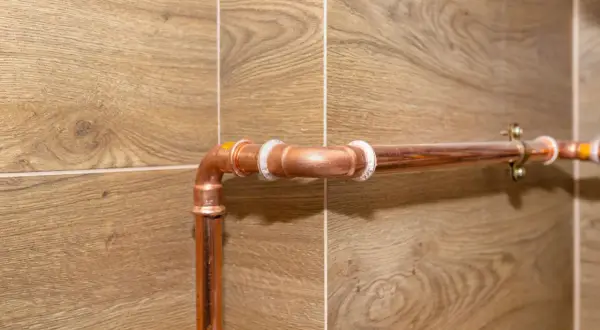 Miedziane rury na drewnopodobnej ścianie. Zastanawiasz się, jak zamaskować rury na ścianie? Ten obraz pokazuje instalację, która może być inspiracją.