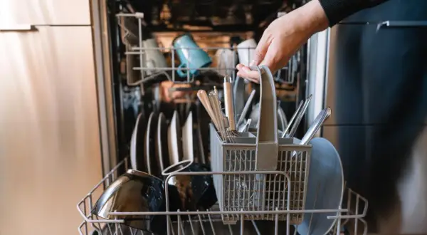 Ręka wkłada koszyk ze sztućcami do zmywarki. To oznacza, że naczynia są "dishwasher safe", czyli można je myć w zmywarce.