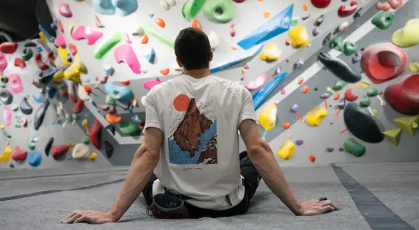 Mann sitzt vor einer Boulderwand mit bunten Griffen. Er ist bereit, besser klettern zu lernen.