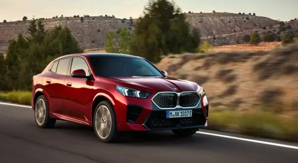 Czerwone BMW X2 2025 pędzi po pustynnej drodze, otoczone suchymi wzgórzami i drzewami.