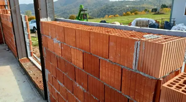 Muro in costruzione con blocchi di laterizio, un materiale che resiste al fuoco, con vista su colline verdi e un escavatore.