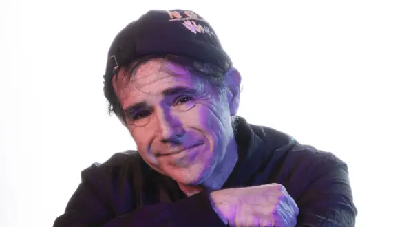 Andrea Satta, con un sorriso enigmatico, indossa un cappellino scuro e una felpa. La luce colorata crea un'atmosfera suggestiva.
