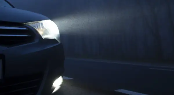 El coche avanza en la niebla con sus luces antiniebla encendidas, iluminando el camino oscuro y los árboles a un lado.