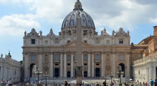 La maestosa facciata di San Pietro domina la piazza, con la sua cupola imponente e l'obelisco centrale. Turisti ammirano la grandezza.