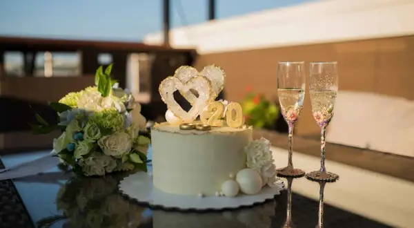 Torta per 20 anni di matrimonio con cuori, anelli e numero 20 dorato. Due flute di champagne decorate e un bouquet di fiori completano la scena.