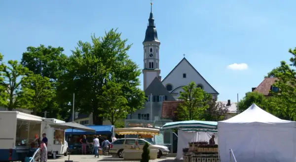 Bunter Markt in Mering mit Kirche im Hintergrund. Stände mit Blumen und Lebensmitteln, Menschen bummeln.