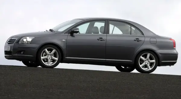 Szary sedan Toyota Avensis 1.8 benzyna stoi na ciemnej nawierzchni, z niebieskim niebem w tle.