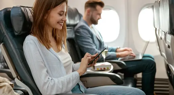 Kobieta i mężczyzna w samolocie. Ona korzysta z telefonu, on pracuje na laptopie. Tak działa tryb samolotowy, pozwalając na używanie urządzeń elektronicznych.
