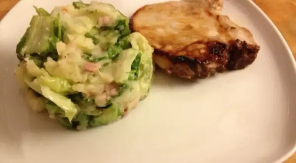 Ein gebratenes Schweinekotelett liegt neben einem Berg aus Kartoffelpüree mit Speck und **endivien untereinander**.