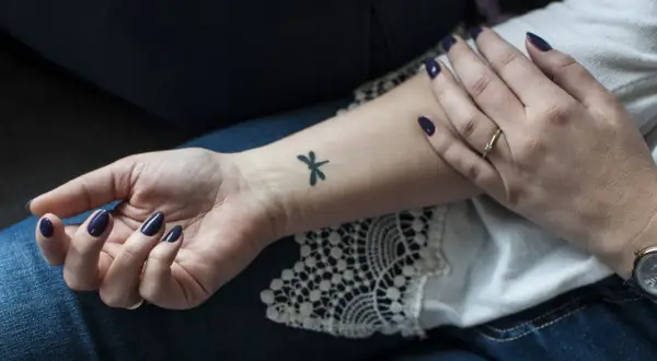 Delikatne, czarne tatuaze male na nadgarstku, w tym ważka. Dłoń ozdobiona ciemnym lakierem i pierścionkiem.