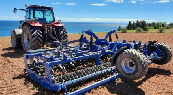 Czerwony traktor z niebieskim agregatem uprawowym na polu, gotowy do siewu. W tle błękitne niebo i morze.
