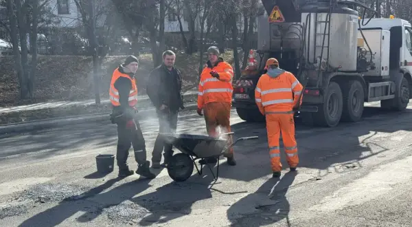Ekipa drogowców w pomarańczowych strojach pracuje przy drodze, obok wózka i specjalistycznej ciężarówki.